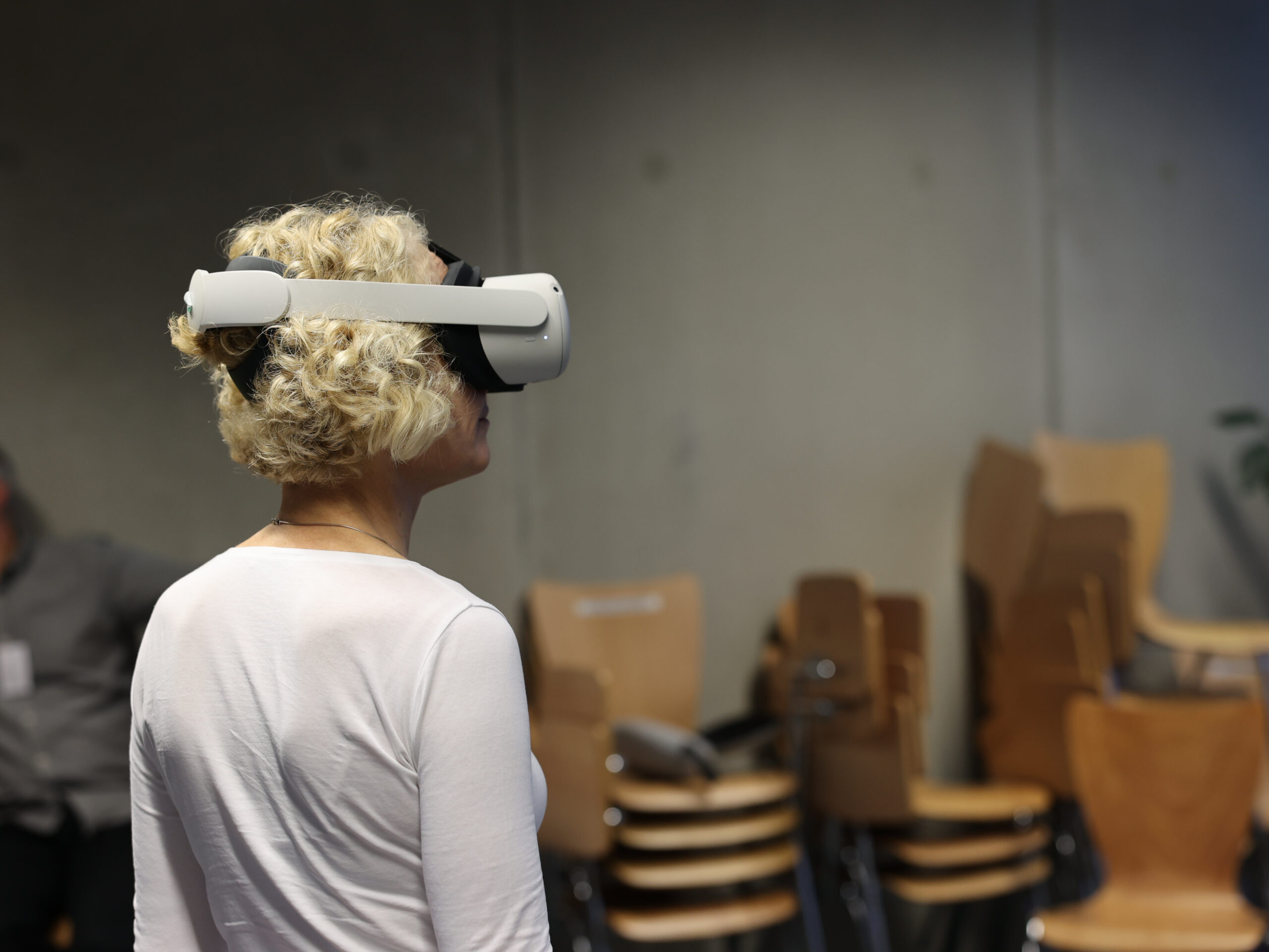 Eine Frau mit lockigem blondem Haar trägt ein VR-Headset und blickt in die Ferne. Im Hintergrund sind leere Stühle und verschwommen erkennbare Personen in einem Raum zu sehen.