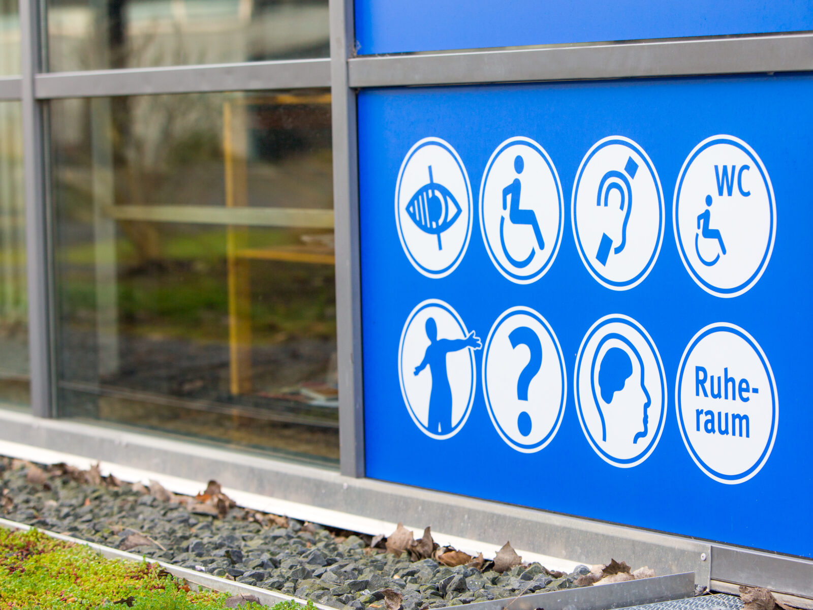 Das Bild zeigt eine blaue Gebäudewand mit acht weißen Piktogrammen, die verschiedene barrierefreie Angebote darstellen. Zu sehen sind Symbole für Unterstützung bei Seh- und Hörbeeinträchtigungen, ein rollstuhlgerechtes WC, einen Ruheraum, einen Informationspunkt sowie weitere Orientierungshilfen.