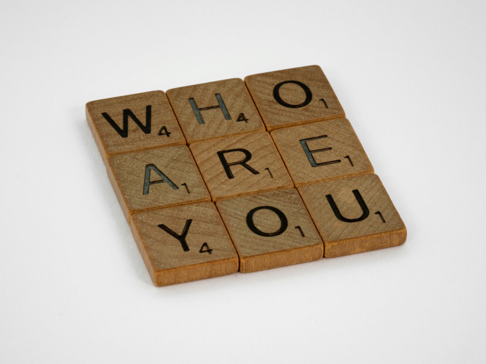 Das Bild zeigt neun Spielsteine aus einem Scrabble-ähnlichen Spiel, die in einem 3x3-Raster angeordnet sind und die Frage „WHO ARE YOU?“ (Wer bist du?) bilden. Die Steine sind aus hellem Holz und die Buchstaben sind schwarz aufgedruckt. Der Hintergrund ist weiß.