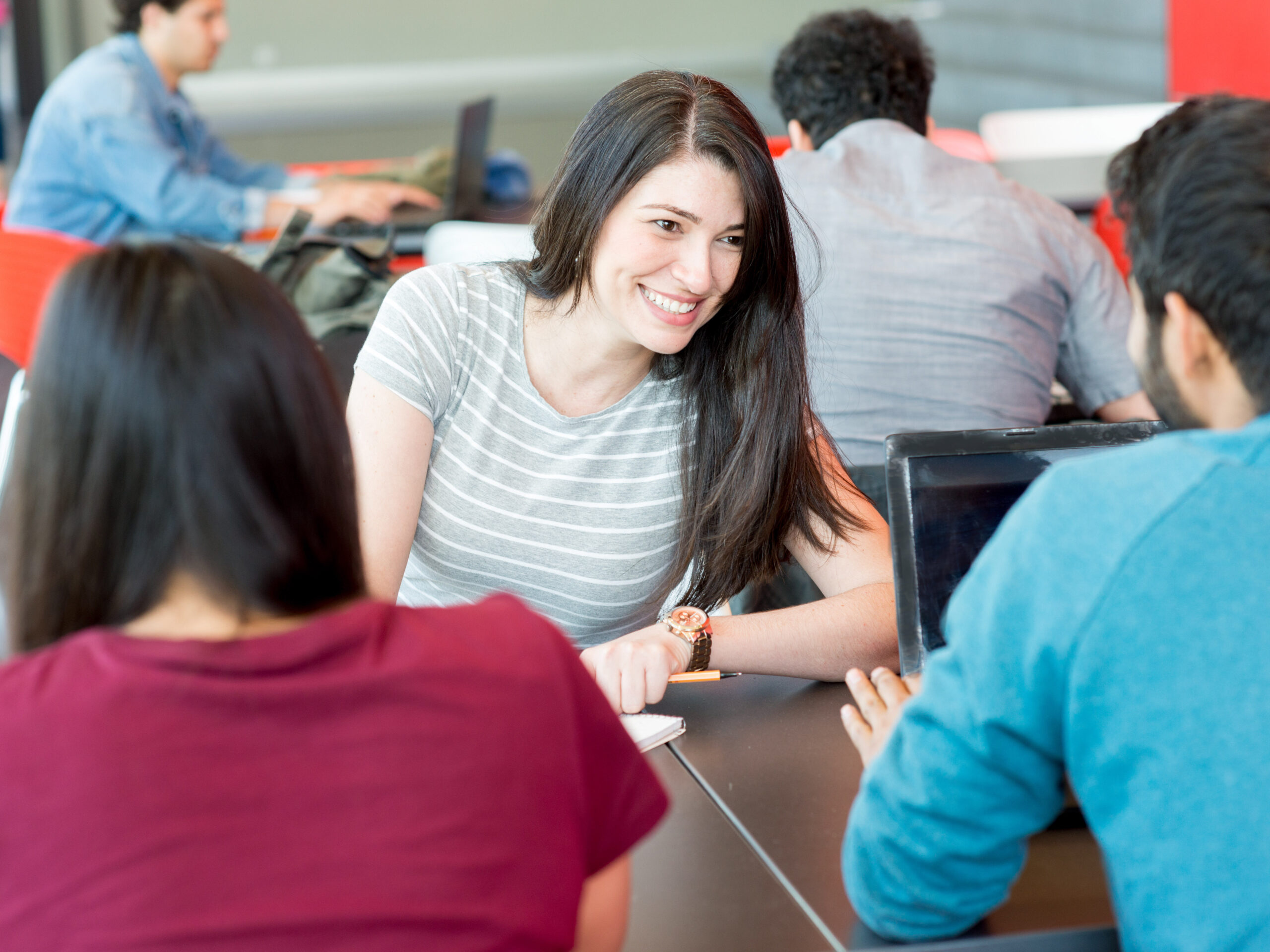 Das Bild zeigt eine Gruppe von Menschen, die sich an einem Tisch treffen. Sie scheinen sich zu unterhalten und zu diskutieren. Im Hintergrund sind weitere Personen zu sehen, die an Computern arbeiten.