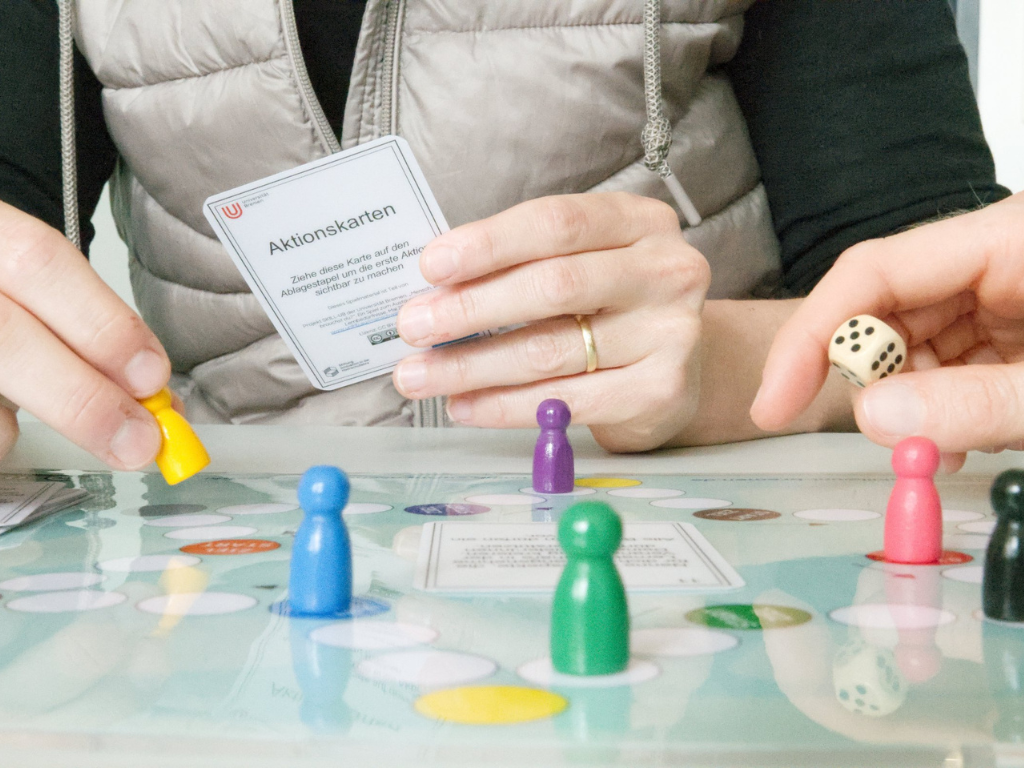 The picture shows a person playing a board game. She is holding an action card in her hands, while colored game pieces and dice are scattered across the game board.