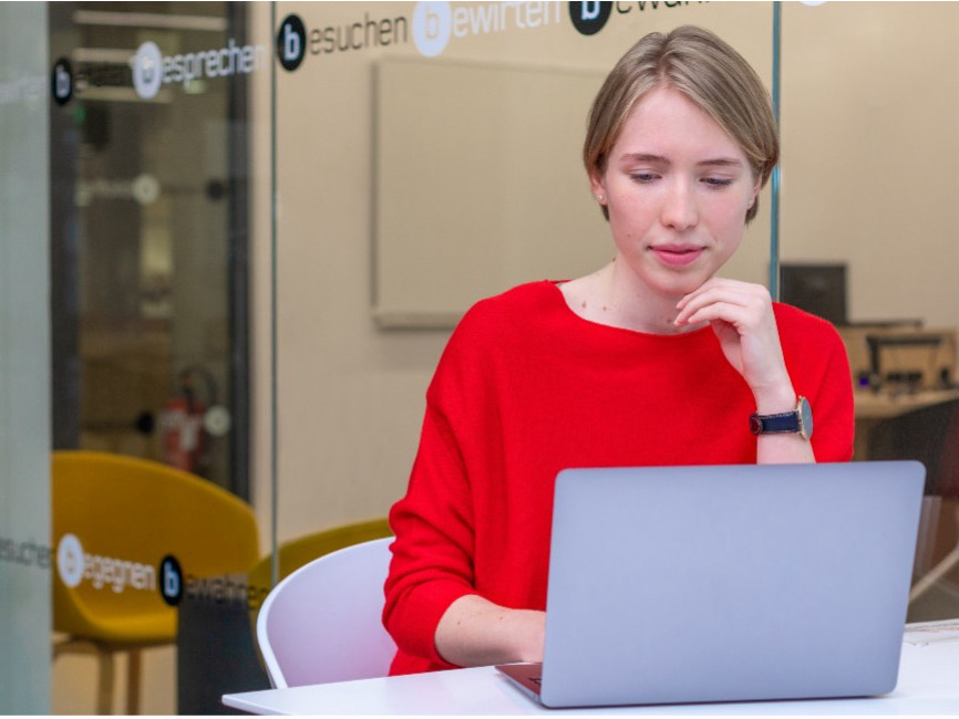 Eine junge Frau mit blonden Haaren, gekleidet in Rot, sitzt vor einem Laptop und arbeitet konzentriert.