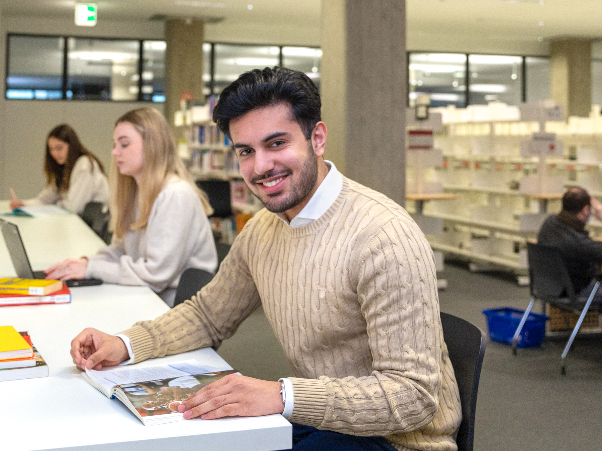 Ein lächelnder junger Mann sitzt an einem Tisch in einer Bibliothek und liest ein Buch. Im Hintergrund sind weitere Personen und Bücherregale zu sehen.
