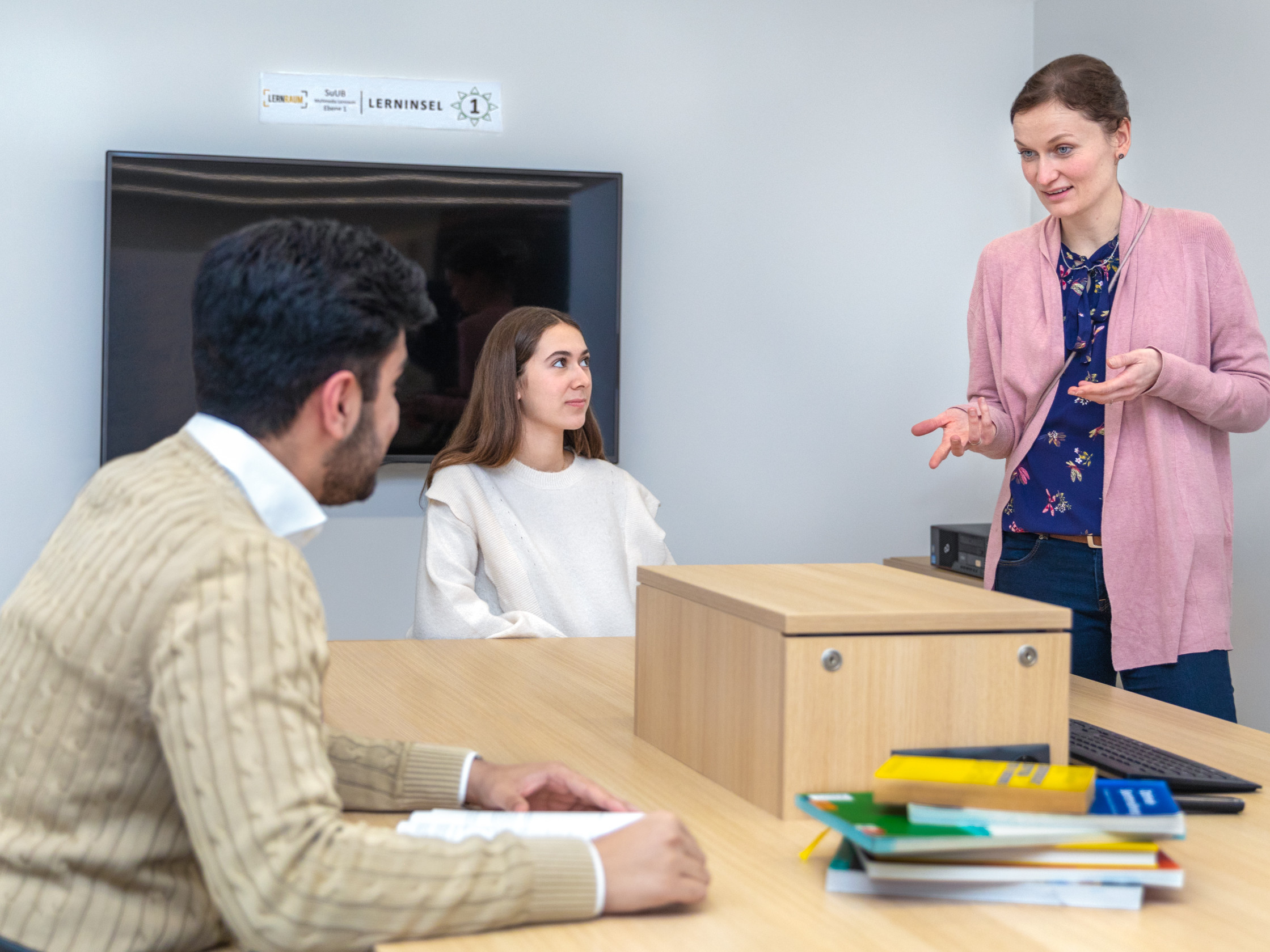 Das Bild zeigt eine Frau, die vor einer kleinen Gruppe von Studenten spricht und gestikuliert. Sie steht an einem Tisch in einem modernen Lernraum, während die Studenten ihr aufmerksam zuhören.