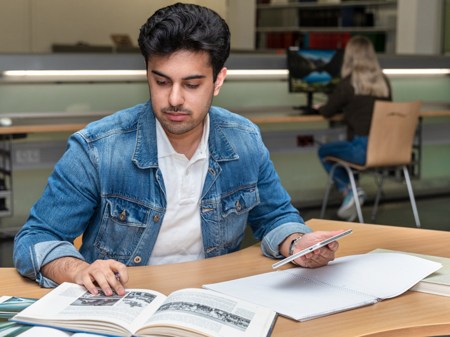 Ein junger Mann mit dunklen Haaren und einer Jeansjacke sitzt an einem Holztisch in einer Bibliothek. Er hält einen Stift in der Hand und schreibt in ein kariertes Notizbuch, während er auf ein aufgeschlagenes Buch mit Bildern blickt.