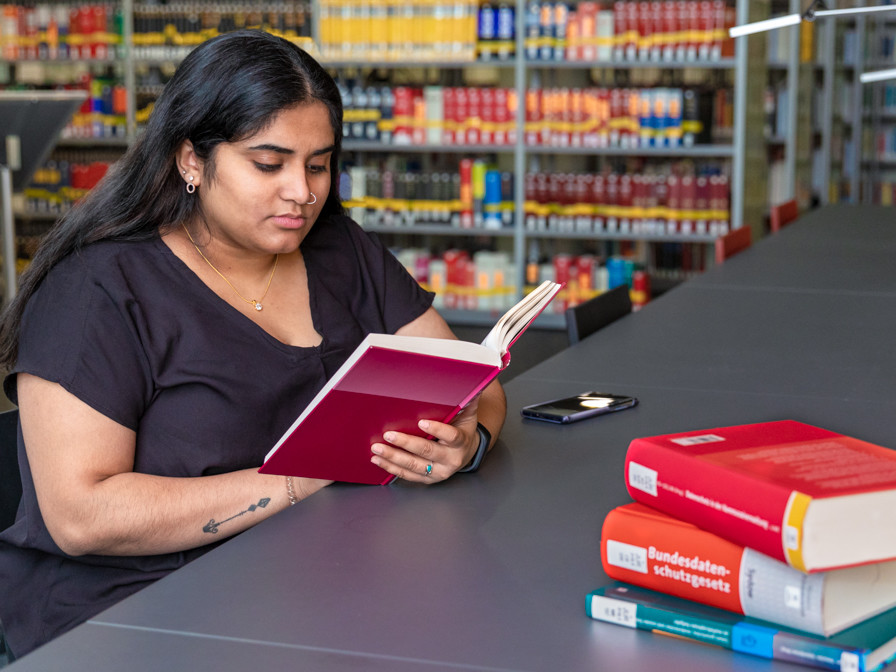 Eine junge Frau mit langen, dunklen Haaren liest konzentriert ein rotes Buch an einem Tisch in einer Bibliothek, umgeben von Bücherregalen.