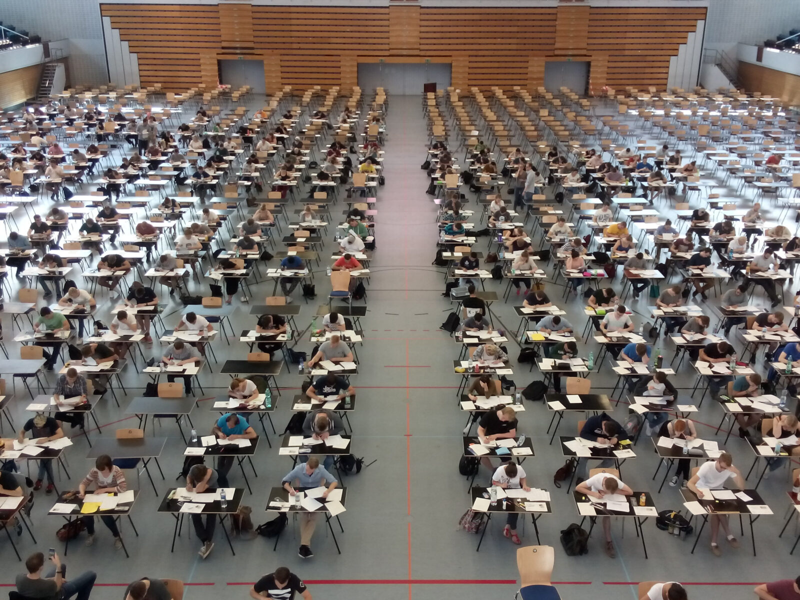 Das Bild zeigt eine große Halle, die für eine Prüfung eingerichtet wurde, mit zahlreichen Schreibtischen in Reihen angeordnet. Viele Personen sitzen an den Tischen und bearbeiten Prüfungsaufgaben, die vor ihnen liegen. Die Halle ist mit einer blauen Linie in zwei Bereiche unterteilt.