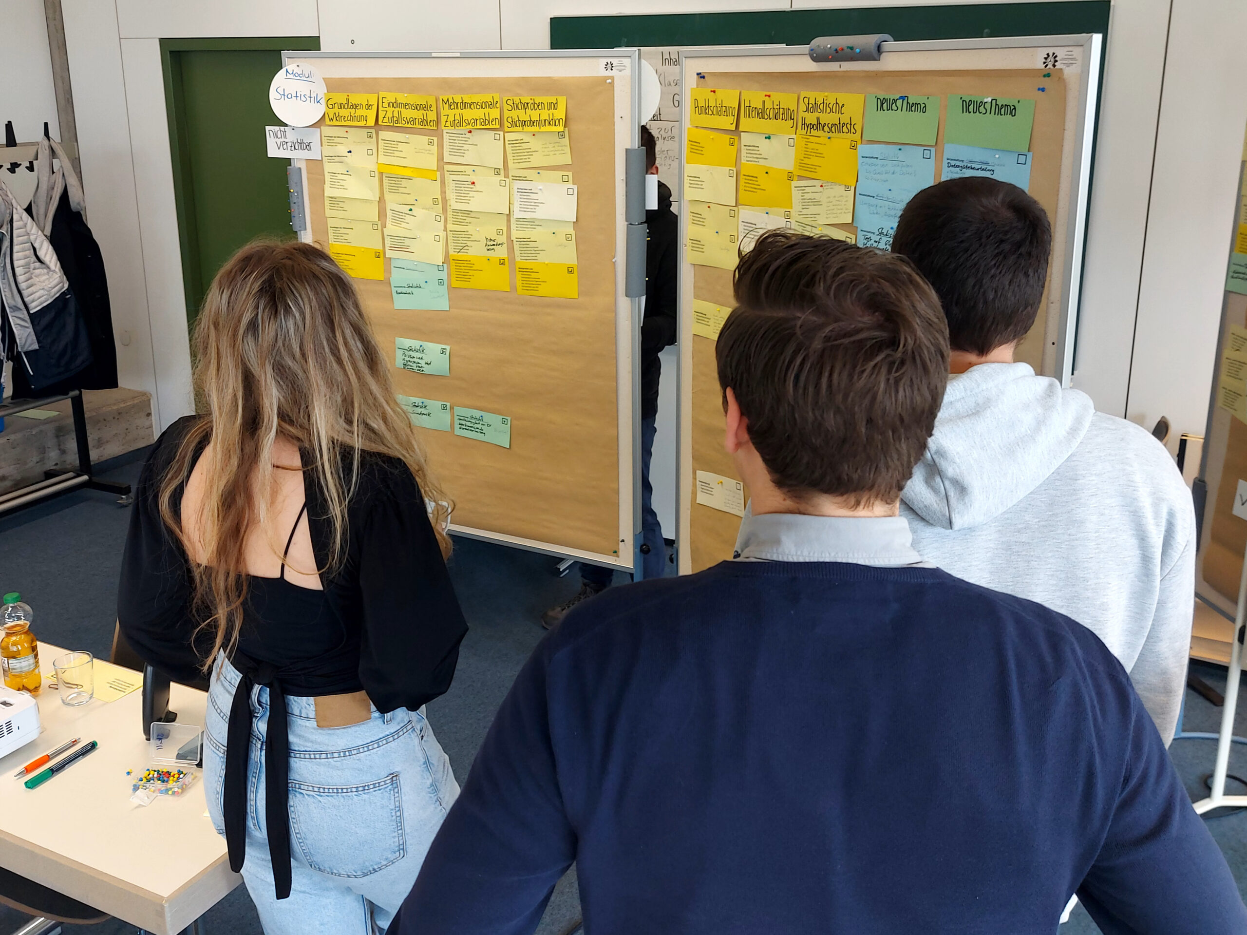 Drei Personen stehen vor zwei Pinnwänden in einem Seminarraum und betrachten Notizzettel. Es handelt sich um eine Workshop-Situation.