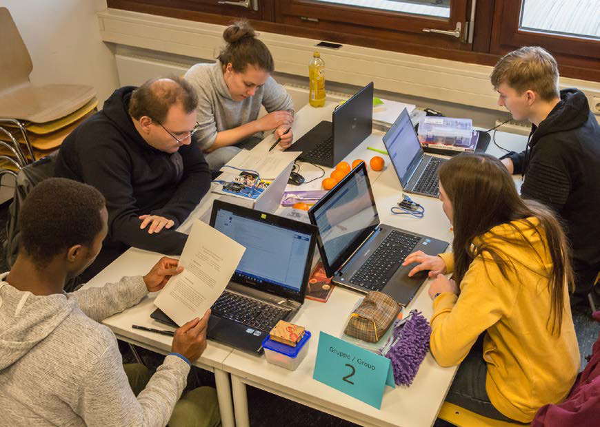 Das Bild zeigt eine Gruppe von vier jungen Leuten, die um einen Tisch sitzen und an Laptops arbeiten. Sie scheinen an einem gemeinsamen Projekt zu arbeiten, möglicherweise im Rahmen eines Kurses oder Workshops, wie ein Schild mit der Aufschrift "Gruppe 2" andeutet. Auf dem Tisch befinden sich neben den Laptops auch Notizen, Obst und andere Gegenstände, die auf eine konzentrierte, aber entspannte Arbeitsatmosphäre hindeuten.