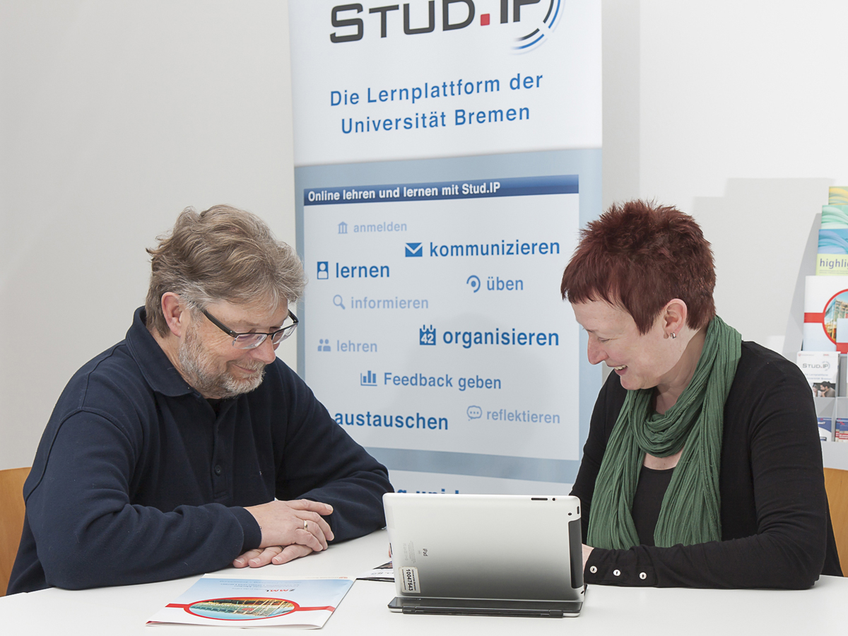 Das Bild zeigt zwei Personen, die an einem Tisch sitzen und sich unterhalten. Im Hintergrund befindet sich ein Roll-up-Banner mit dem Logo und Informationen zu "Stud.IP", einer Lernplattform der Universität Bremen. Die Szene deutet auf eine Beratung oder einen Austausch bezüglich der Nutzung der Lernplattform hin.