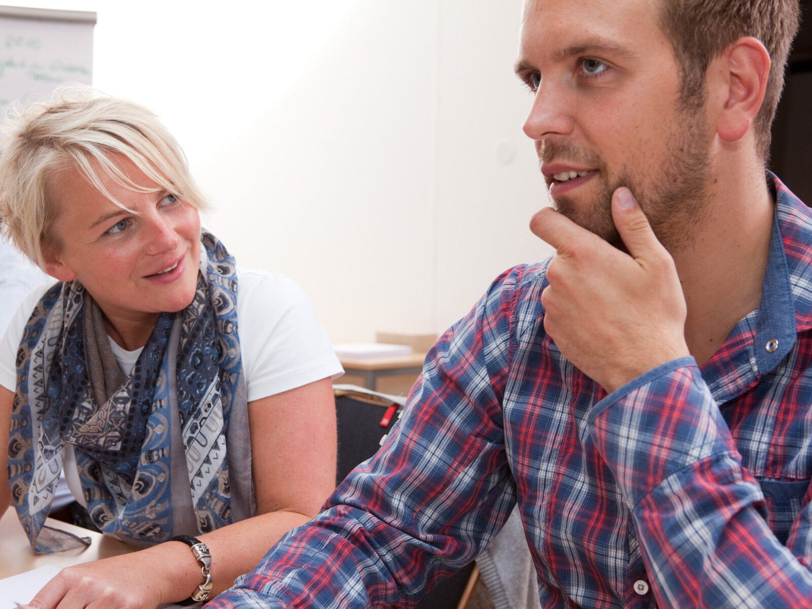 Ein Mann und eine Frau sitzen an einem Tisch nebeneinander und unterhalten sich angeregt. Der Mann, mit Bart und kariertem Hemd, blickt der Frau zu, während diese lächelt und ihn ansieht.